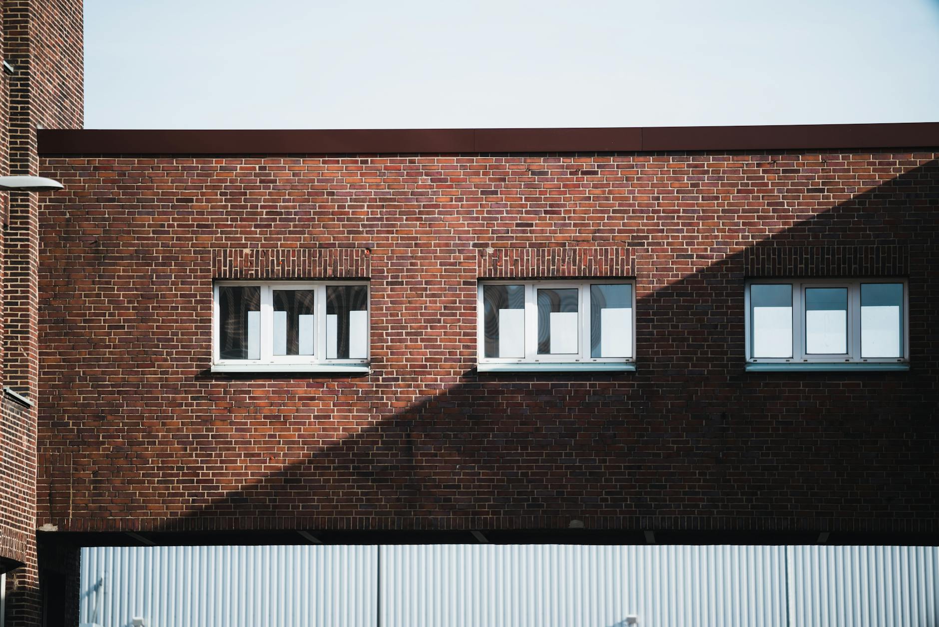 Front view of a red brick building facade with three white windows casting shadows.