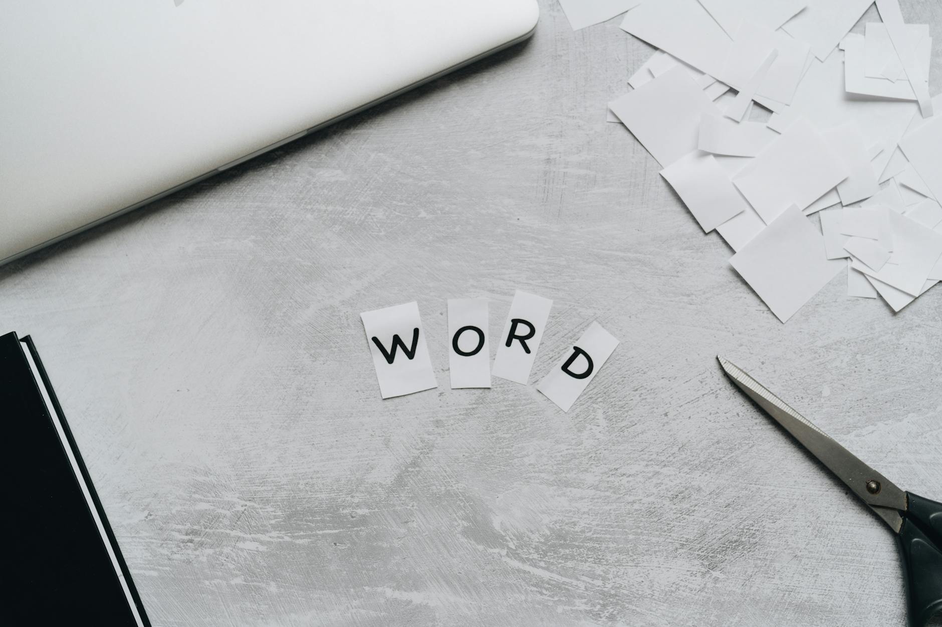 Top view of a workspace with scattered paper letters spelling 'WORD', next to a laptop and scissors.