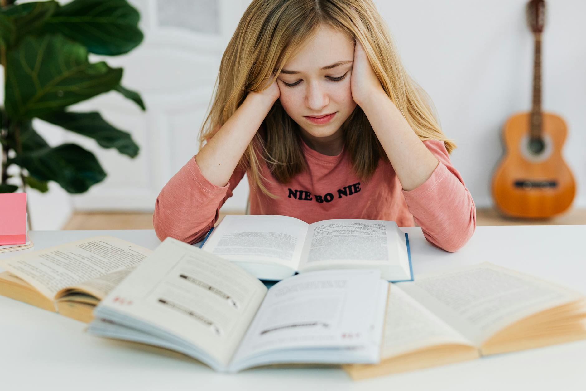 A young woman in a pink sweater focused on reading books at a table indoors.
