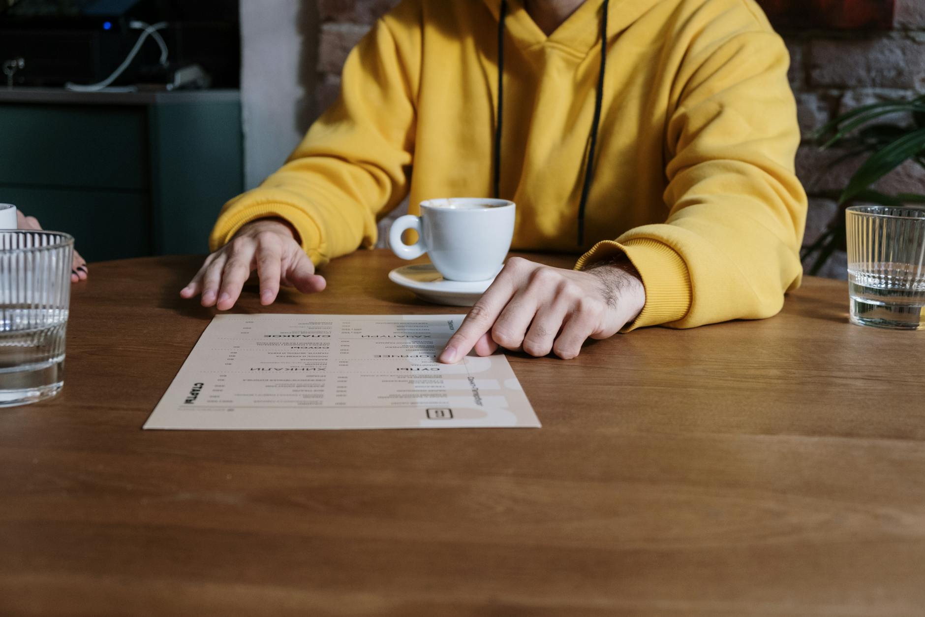 A person wearing a yellow hoodie points to a menu in a cozy cafe setting.