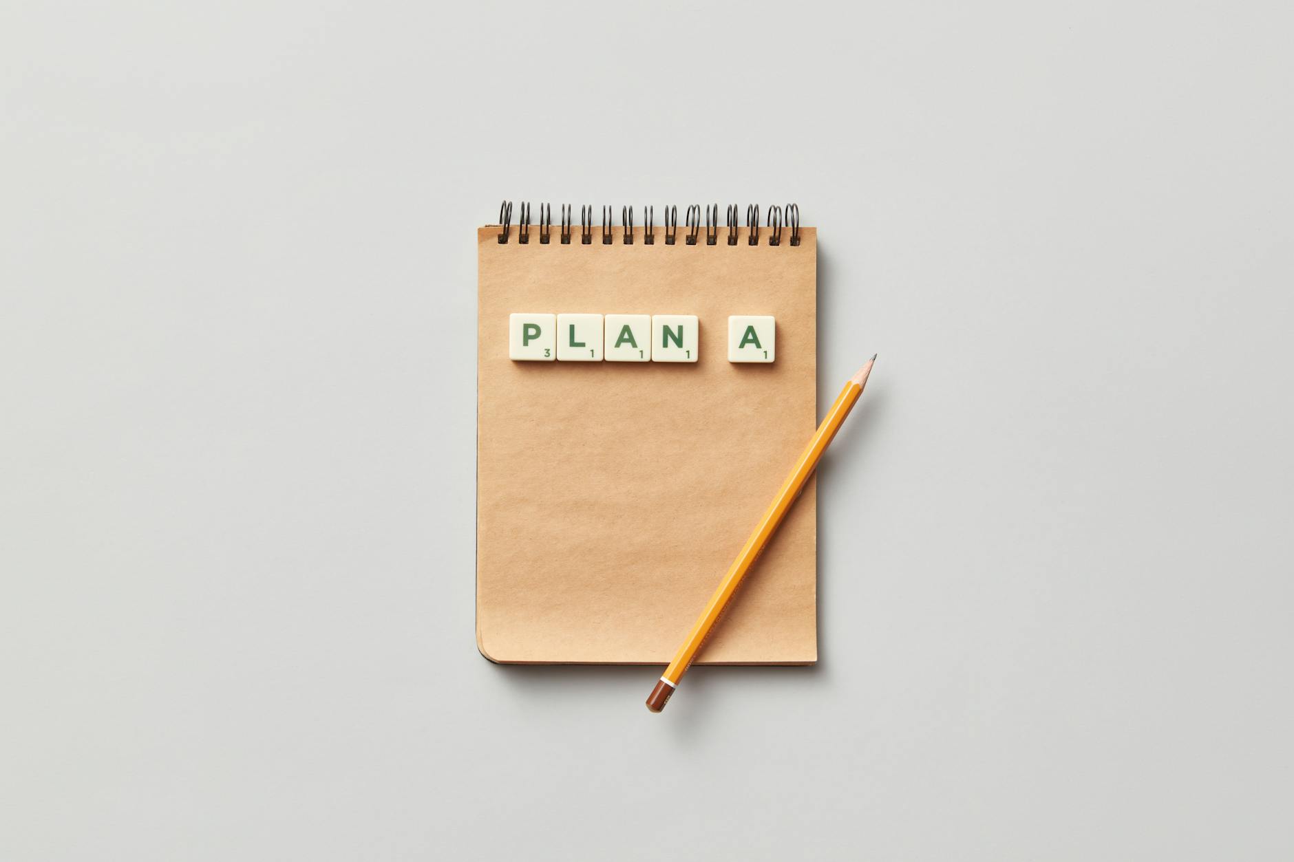 Top view of a notepad with a pencil and scrabble tiles spelling 'PLAN A' on a gray background.