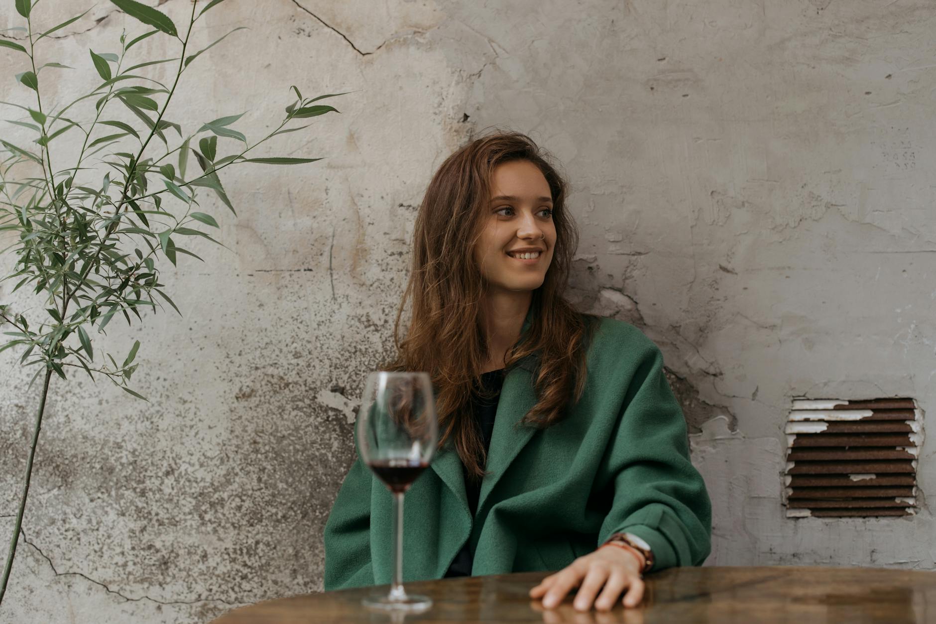 A woman sitting outdoors, smiling with a glass of red wine in hand, wearing a green coat.