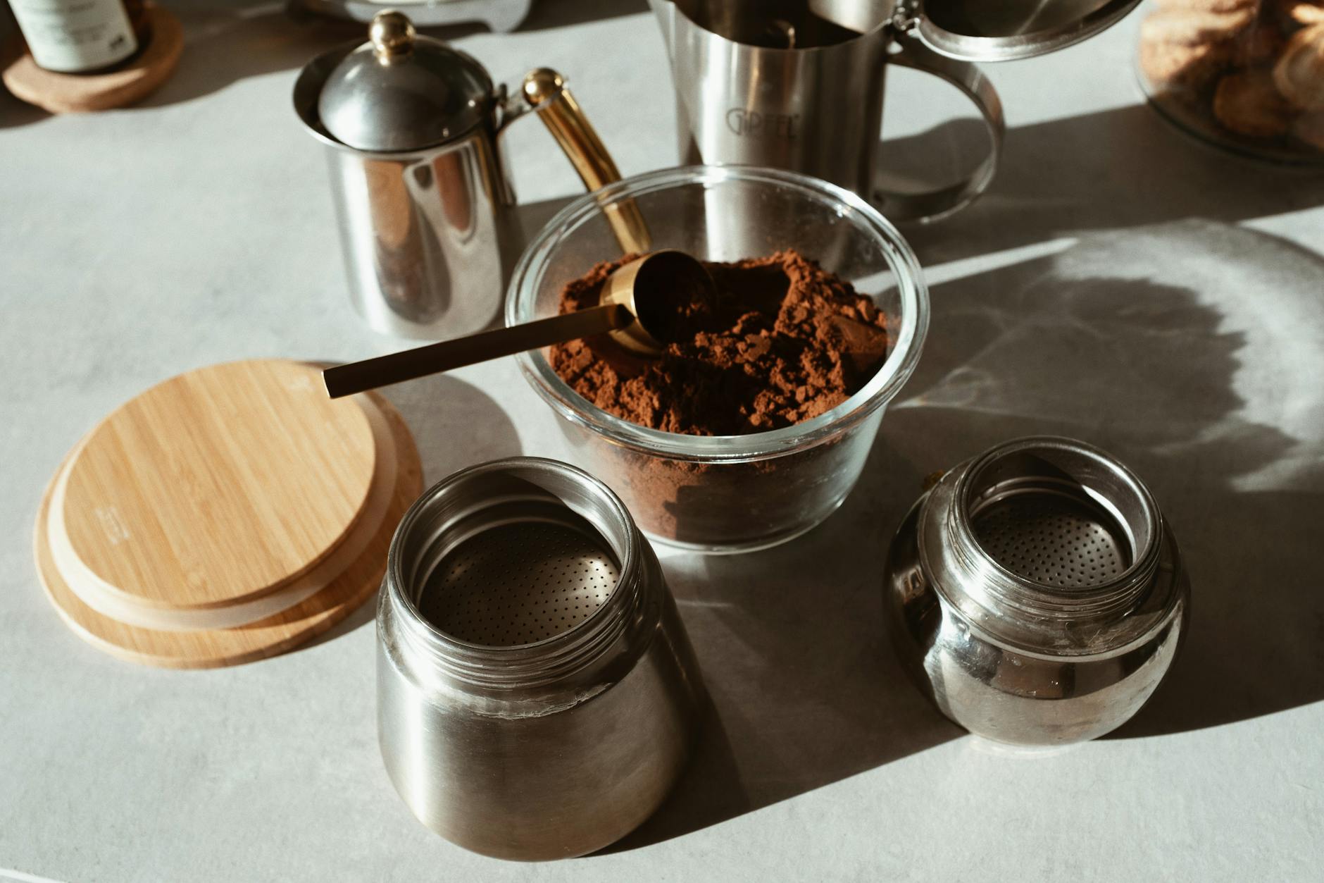 A coffee-making setup with powder in a glass jar, stainless steel containers, and wooden lid.